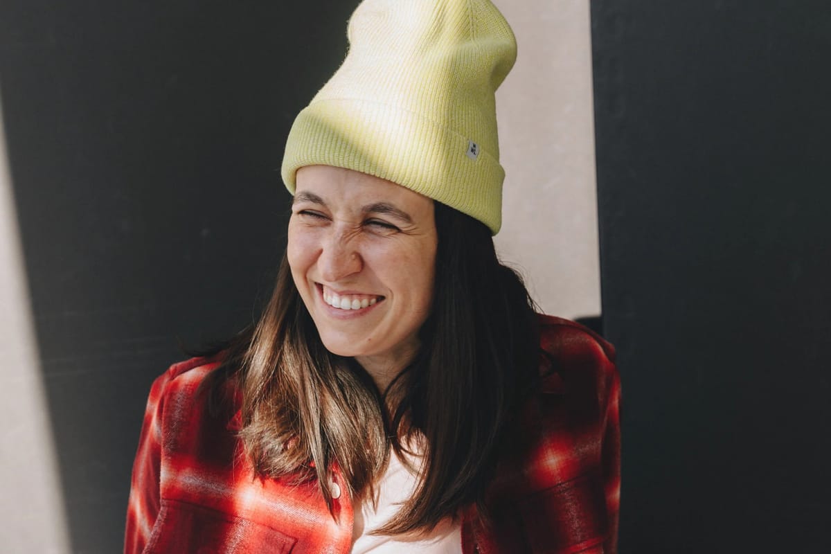 Comedian Ashley Gavin laughing in a yellow beanie and plaid shirt, official press photo