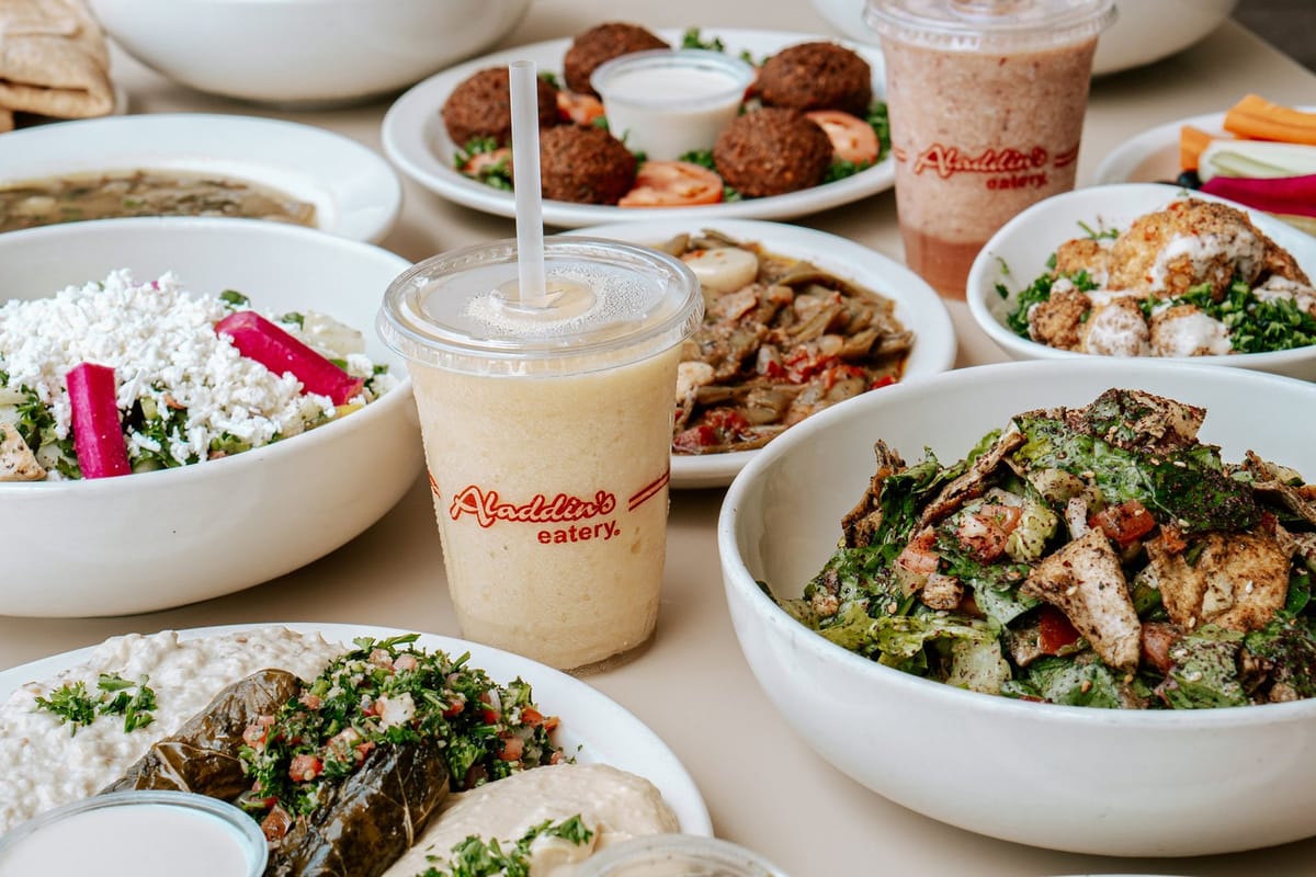 Table spread of Aladdin's Eatery dishes including falafel, fattoush salad, tabouli, stuffed grape leaves, green beans and smoothies in branded cups