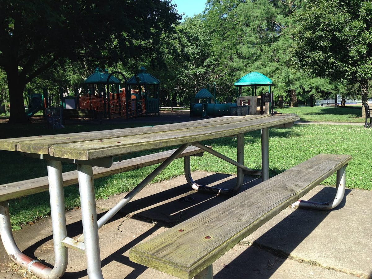 A weathered wooden picnic table on a concrete pad in the foreground, with a playground of green-roofed towers and orange panels under tall trees behind it