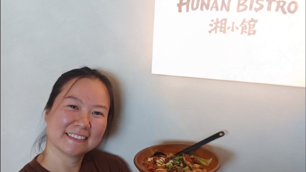 Hunan Bistro co-owner holding a bowl of braised beef noodles beside the lit restaurant sign