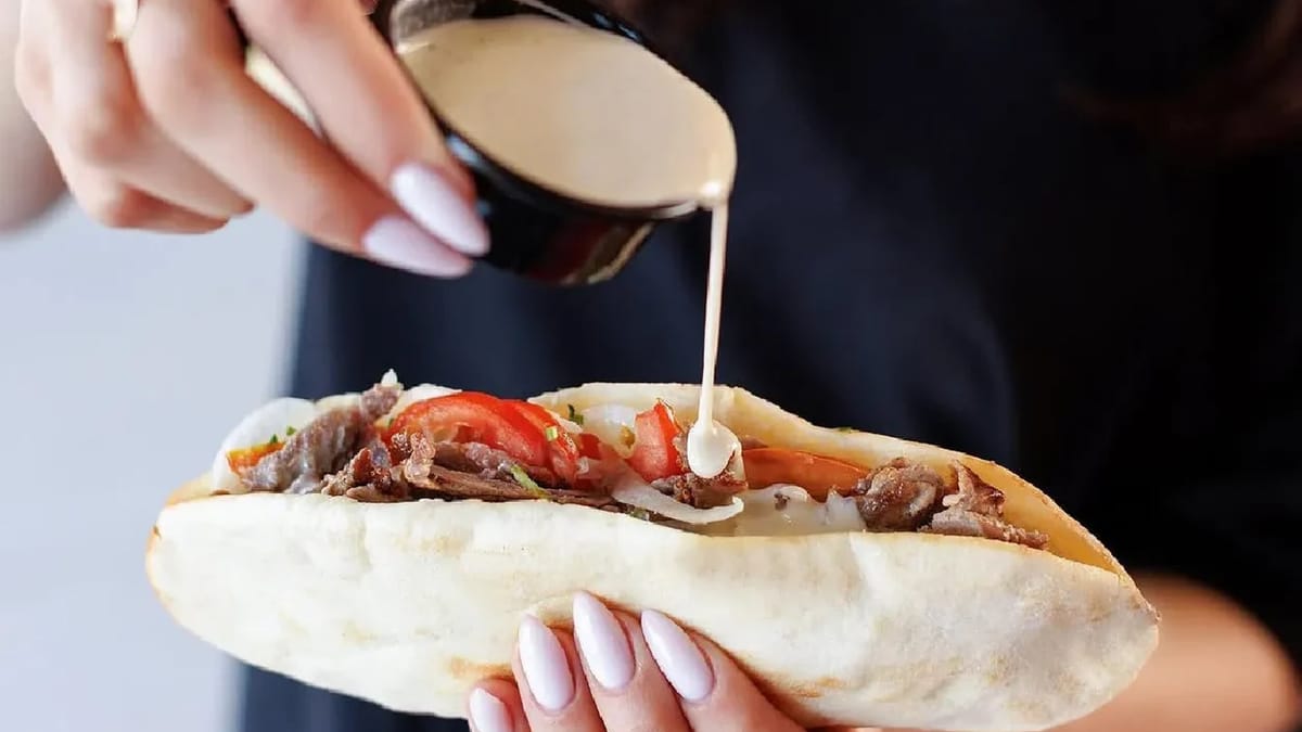 Hands holding a Nashmi Jordanian shawarma sandwich in soft pita while pouring creamy tahini sauce over meat, tomato, and onion