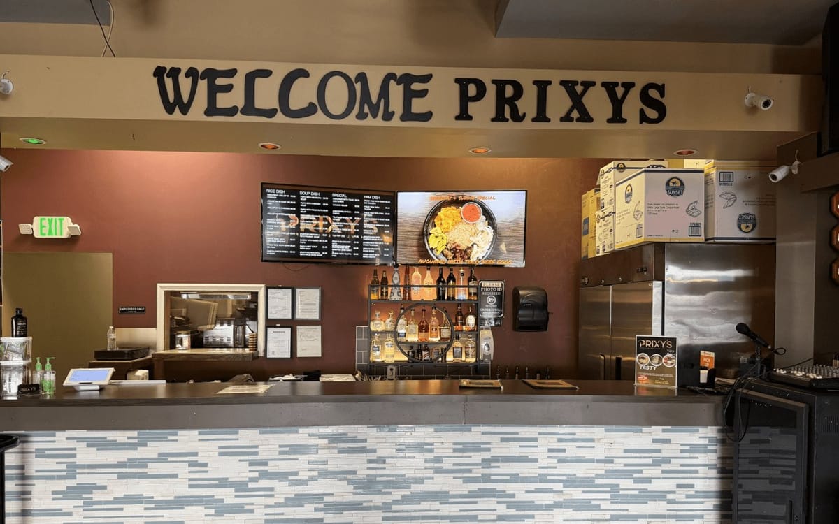 Front counter at Prixys African Restaurant under a Welcome Prixys sign, with digital menu boards and a bottle shelf behind the register