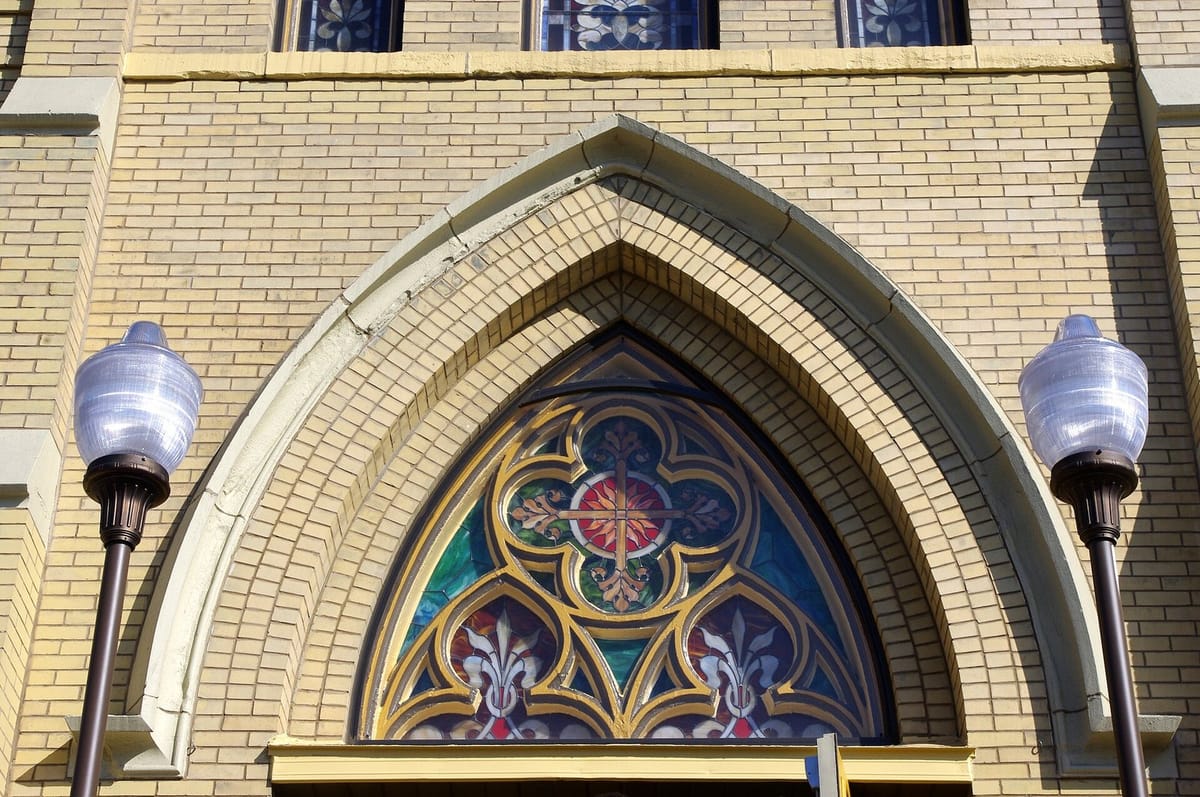 Stained glass window under a pointed brick arch above the front door of Saint John the Baptist Italian Catholic Church, flanked by two street lamps